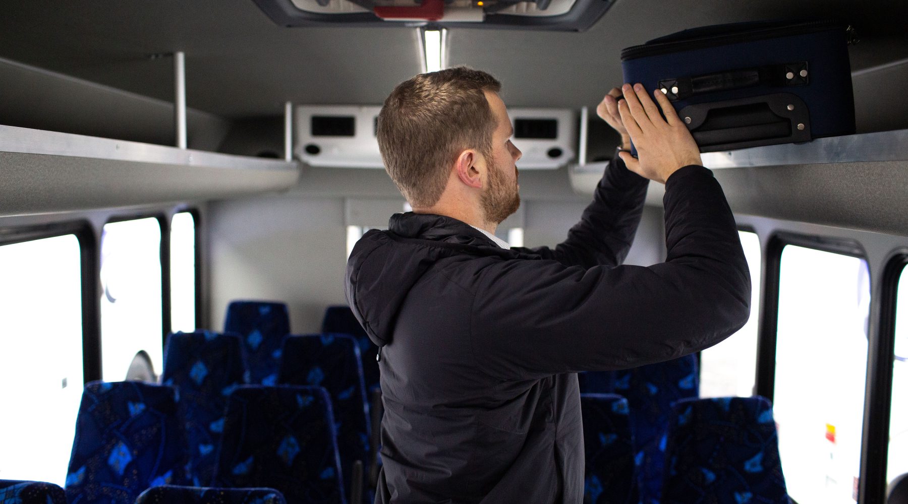 A man puts luggage in storage above a bus seat